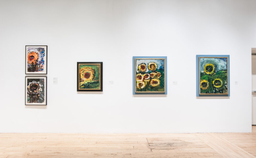 Straight-on view of a white gallery wall featuring five framed sunflower and floral paintings. (Alt-text generated by AI)