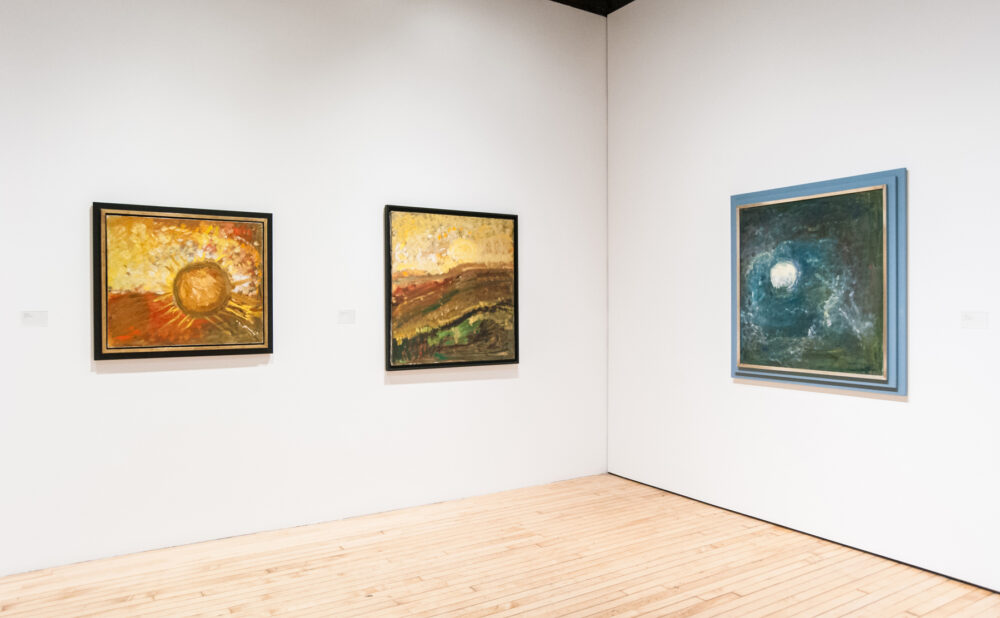 Gallery room corner featuring two small framed paintings on the left wall and a blue-toned painting on the right. (Alt-text generated by AI)