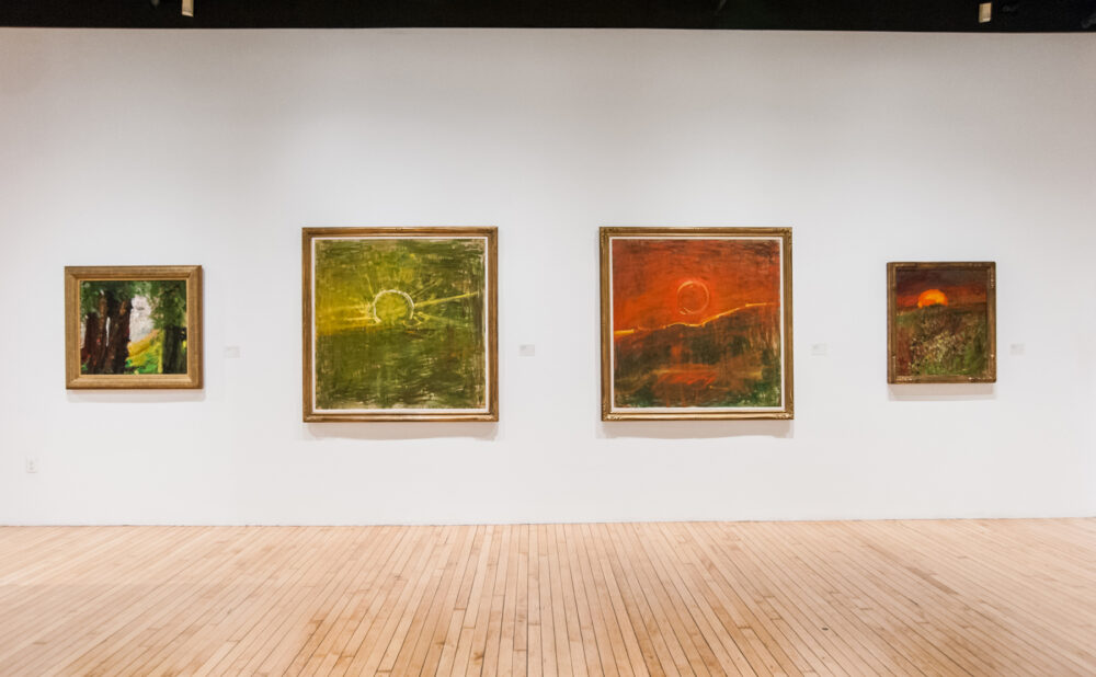 Wide shot of four landscape paintings with vibrant orange and green hues hanging in a row on a white wall. (Alt-text generated by AI)