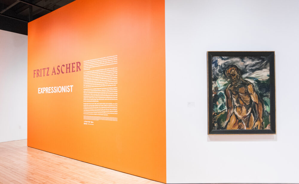 Exhibition entry area featuring a bright orange wall with introductory text on the left and a framed painting on the right. (Alt-text generated by AI)
