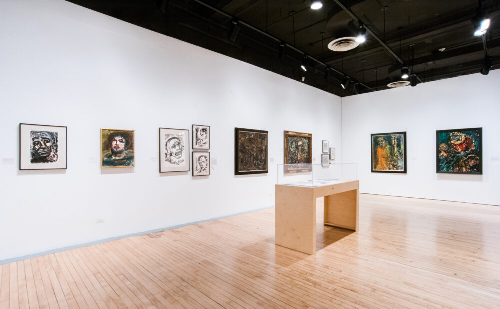 Wide view of a gallery space with dark ceilings, multiple framed works along the walls, and a central display case. (Alt-text generated by AI)