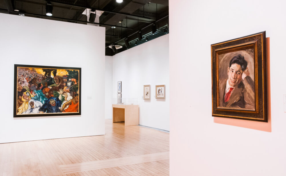 Gallery interior showing a large crowded figure painting on the left and a framed portrait on the right column. (Alt-text generated by AI)