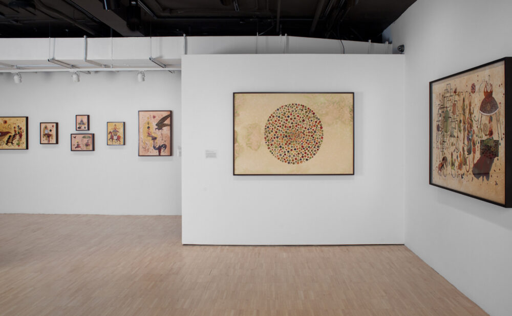 Corner view of the exhibition displaying framed artworks on adjacent white walls, including a large framed drawing on the right. (Alt-text generated by AI)