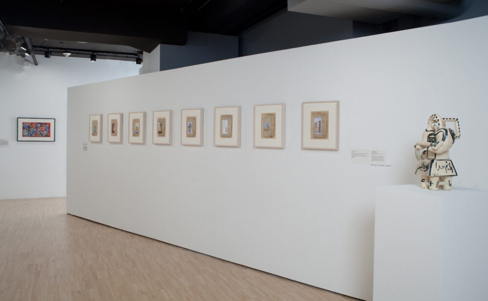 Perspective view along a long white partition wall lined with a series of small, evenly spaced framed artworks. (Alt-text generated by AI)