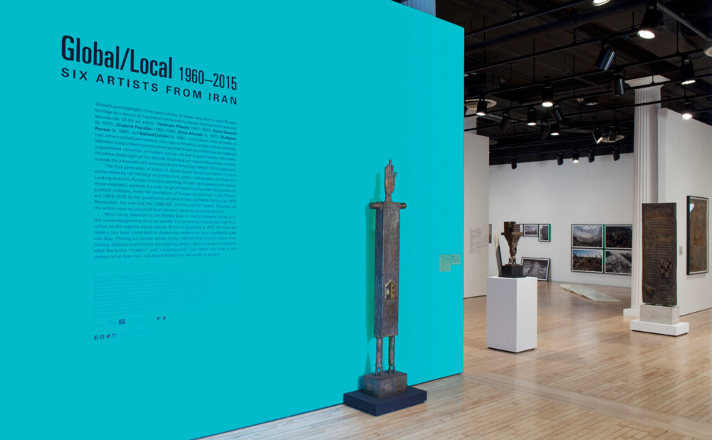 Gallery view featuring a vibrant turquoise accent wall with the title "Global/Local" on it, and an adjacent tall dark sculpture mounted on a plinth. (Alt-text generated by AI)