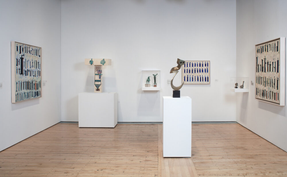 Wide exhibition space showcasing pedestal-mounted sculptures in the center and framed artworks on the surrounding walls. (Alt-text generated by AI)