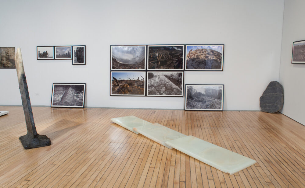 Gallery room displaying photography grids on the wall and a rectangular, light-colored floor-based work on the wooden floor. (Alt-text generated by AI)