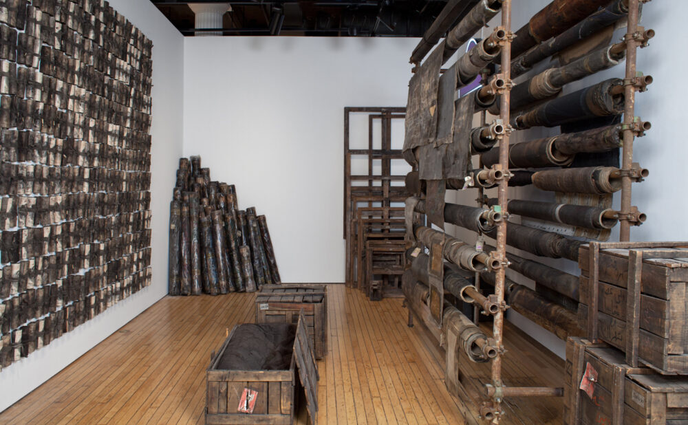 Densely installed gallery area featuring stacked wooden beams, fabric bundles, and a large woven wall hanging. (Alt-text generated by AI)