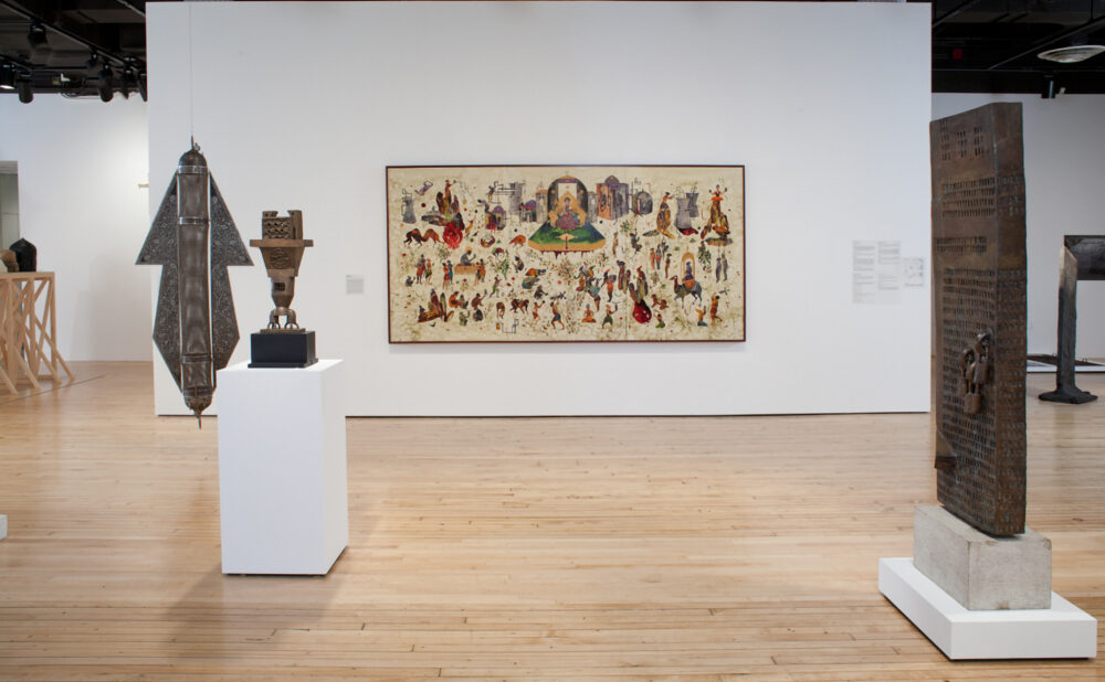 Exhibition view highlighting a central detailed painting flanked by free-standing sculptures on white pedestals. (Alt-text generated by AI)