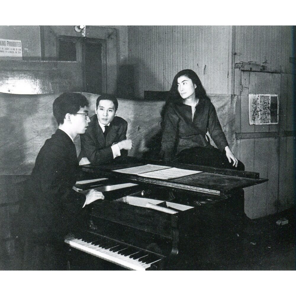 Black-and-white performance photograph showing Yoko Ono at a piano accompanied by musicians in a studio setting. (Alt-text generated by AI)