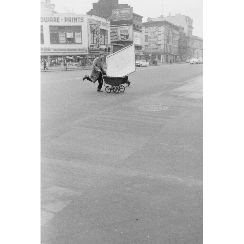 Black-and-white street photograph showing a person pushing a cart down a wide city road. (Alt-text generated by AI)