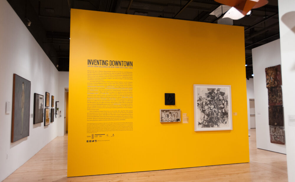 Perspective view of the exhibition entry featuring a bright yellow accent wall printed with the title "Inventing Downtown" and wall text. (Alt-text generated by AI)