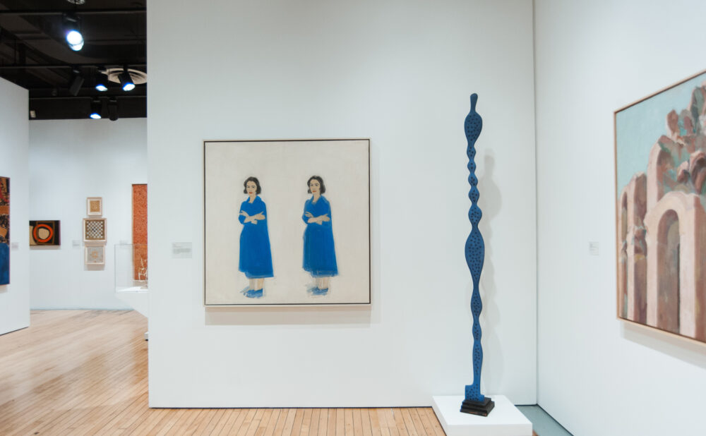 Gallery wall displaying a figurative painting of two figures flanked by a tall, slender blue vertical sculpture. (Alt-text generated by AI)