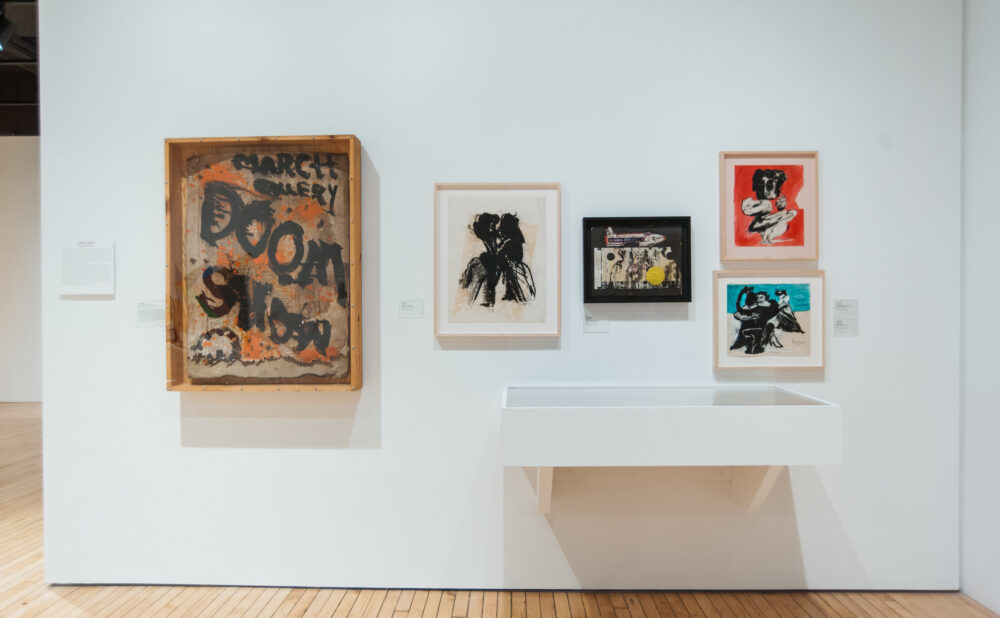 Exhibition wall section featuring framed pop-art posters and graphics above a low white display ledge. (Alt-text generated by AI)