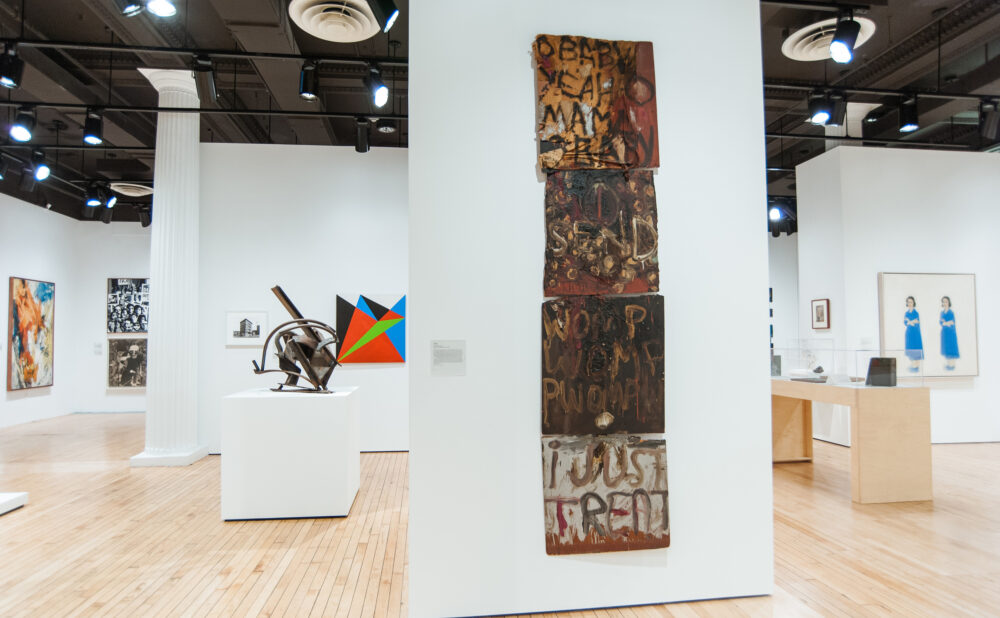 Narrow gallery wall displaying a tall, multi-panel totem-like mixed-media assemblage with sculptures in the background. (Alt-text generated by AI)