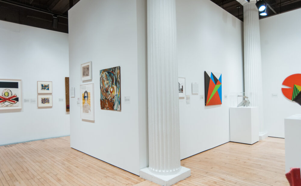 View around a large white architectural column showing surrounding exhibition walls with abstract paintings. (Alt-text generated by AI)