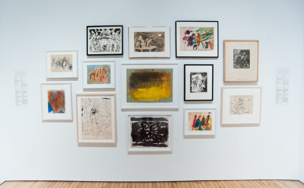 White gallery wall featuring a dense salon-style grid arrangement of small framed drawings and paintings. (Alt-text generated by AI)