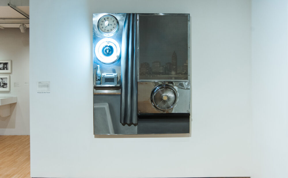 Wall-mounted assemblage work featuring mirrored surfaces, metal fixtures, and fabric elements inside a cutout alcove. (Alt-text generated by AI)