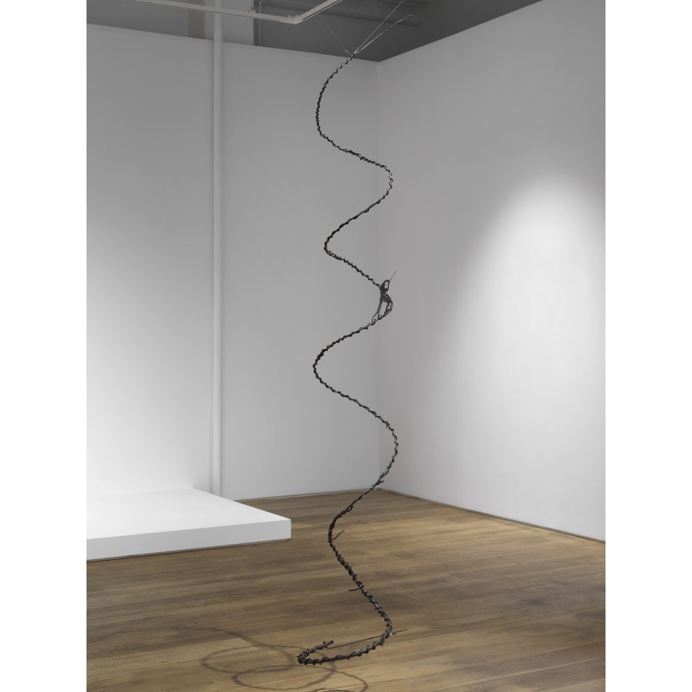 A spiral-shaped black metal sculpture hanging in a minimalist gallery with white walls and a wooden floor.