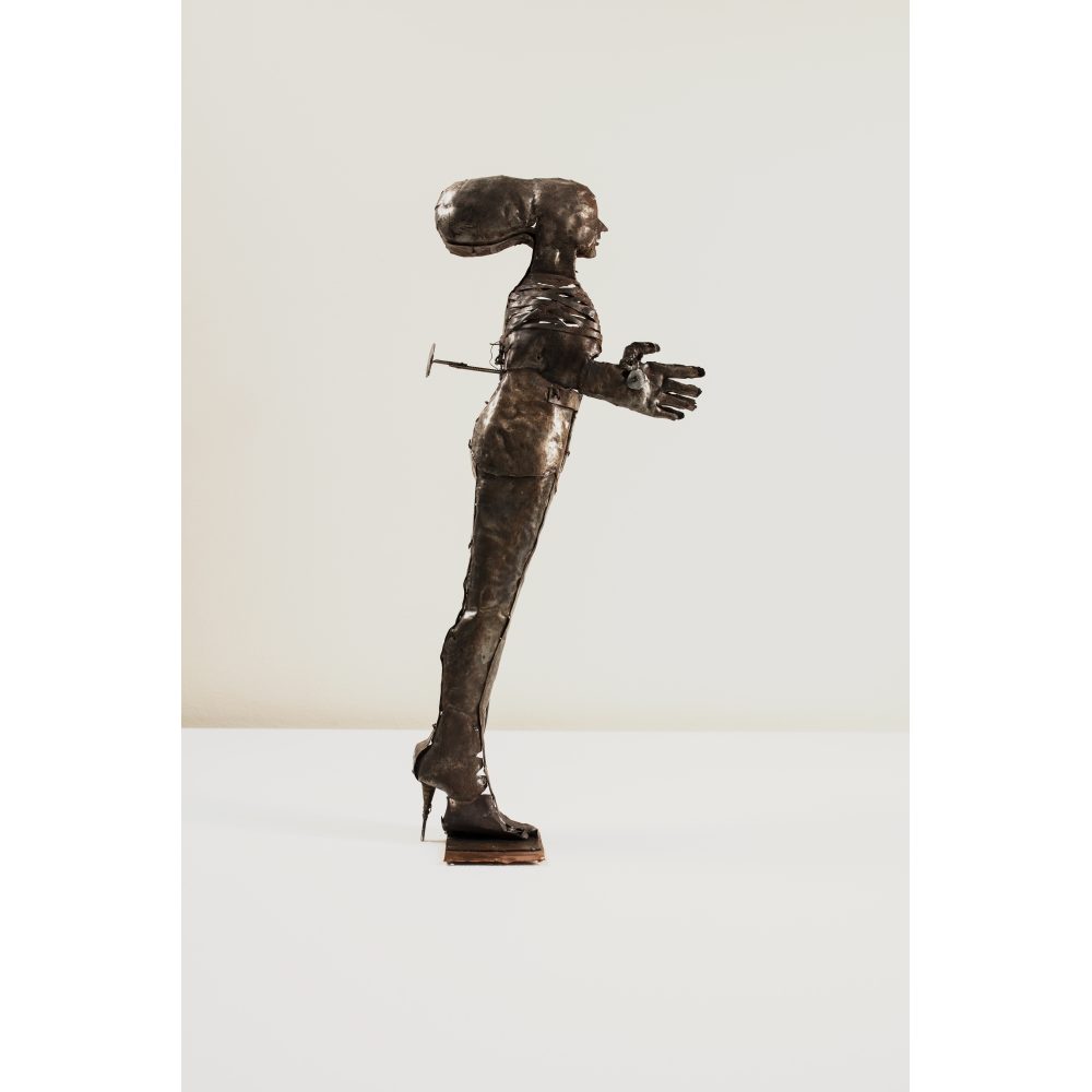 Side profile of a metal humanoid sculpture with an elongated head and exaggerated features, including forward-leaning legs and large hands that reach forward.