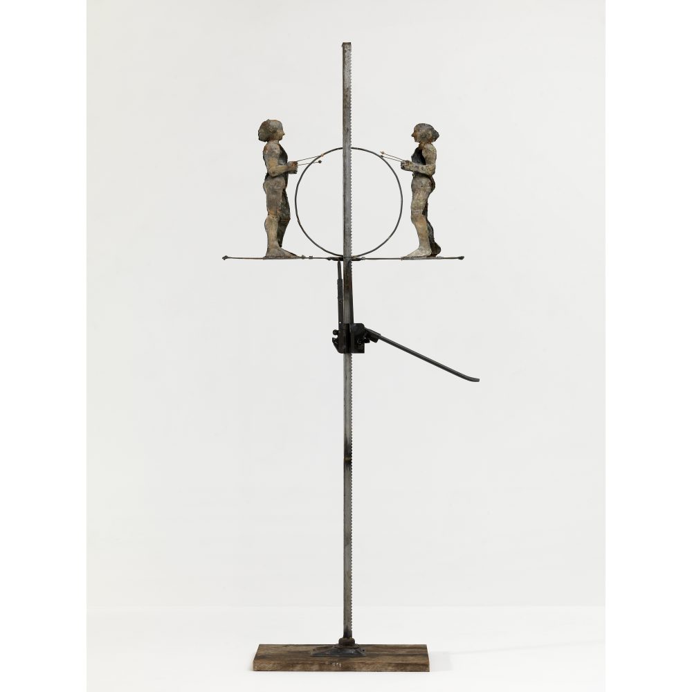 Metal sculpture with two figures facing each other, holding a circular frame, mounted on a vertical pole with a wooden base.