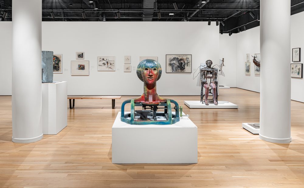 Art gallery with sculptures and paintings on display, featuring at center a colorful head-shaped sculpture on a low white pedestal.