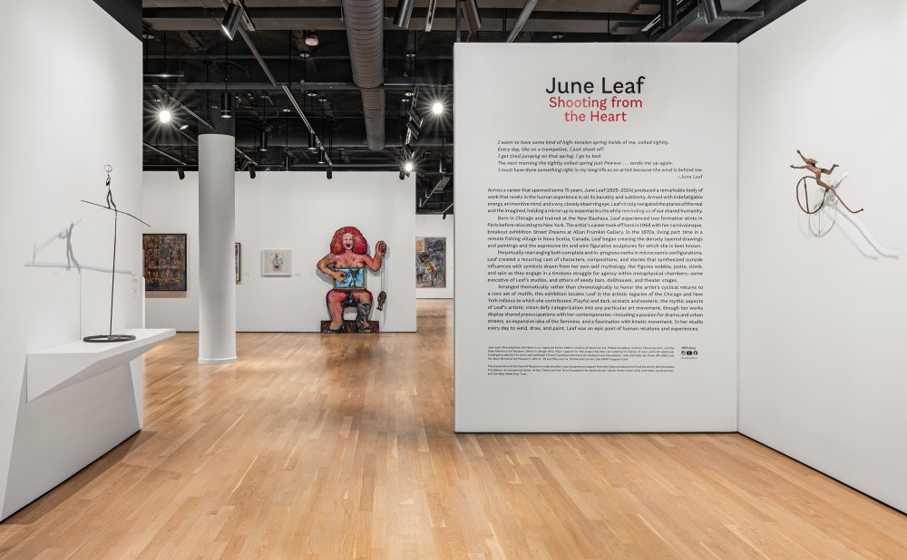 Art gallery interior with sculptures and paintings on display, featuring a text panel titled "June Leaf Shooting from the Heart."
