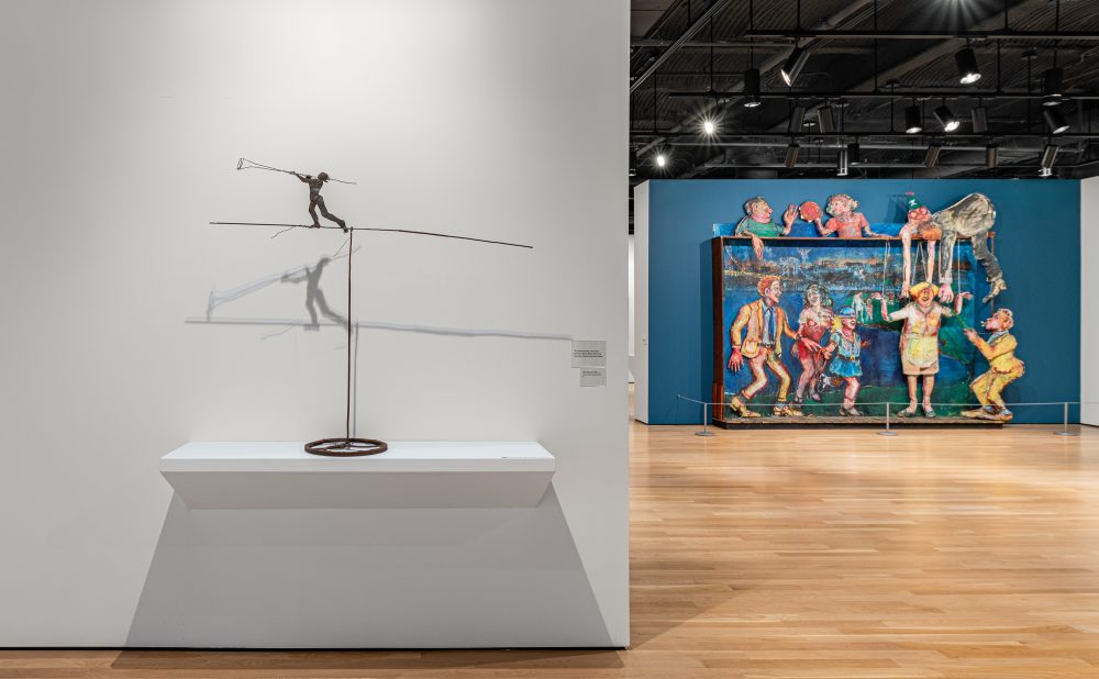 Art gallery with a tightrope walker sculpture and, in the bakcground, a colorful installation of cartoonish figures set against a teal blue wall.