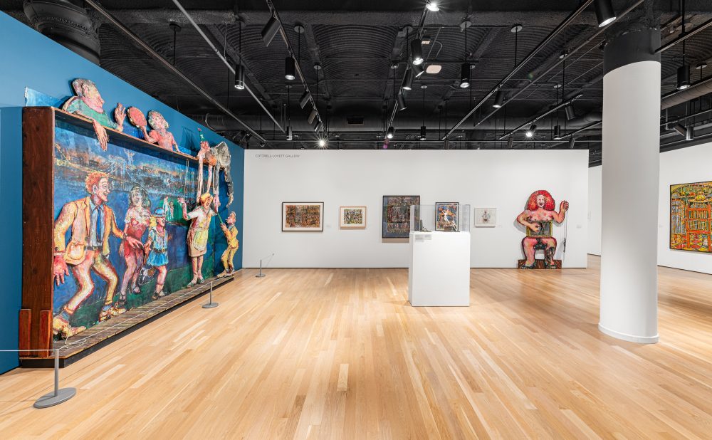 Art gallery with colorful installation against a teal blue wall, a central white pedestal, framed artworks, and red-headed figural sculpture on the wall.