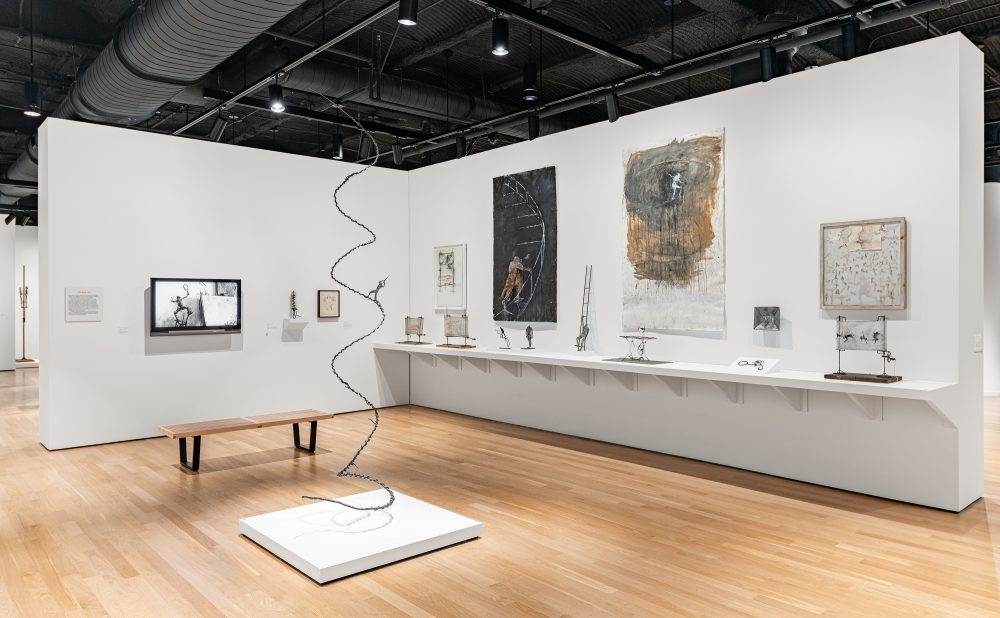Art gallery with sculptures and paintings, featuring a spiral metal sculpture, a bench near a screen displaying a documentary short, and a long, white shelf holding various metal sculptures.