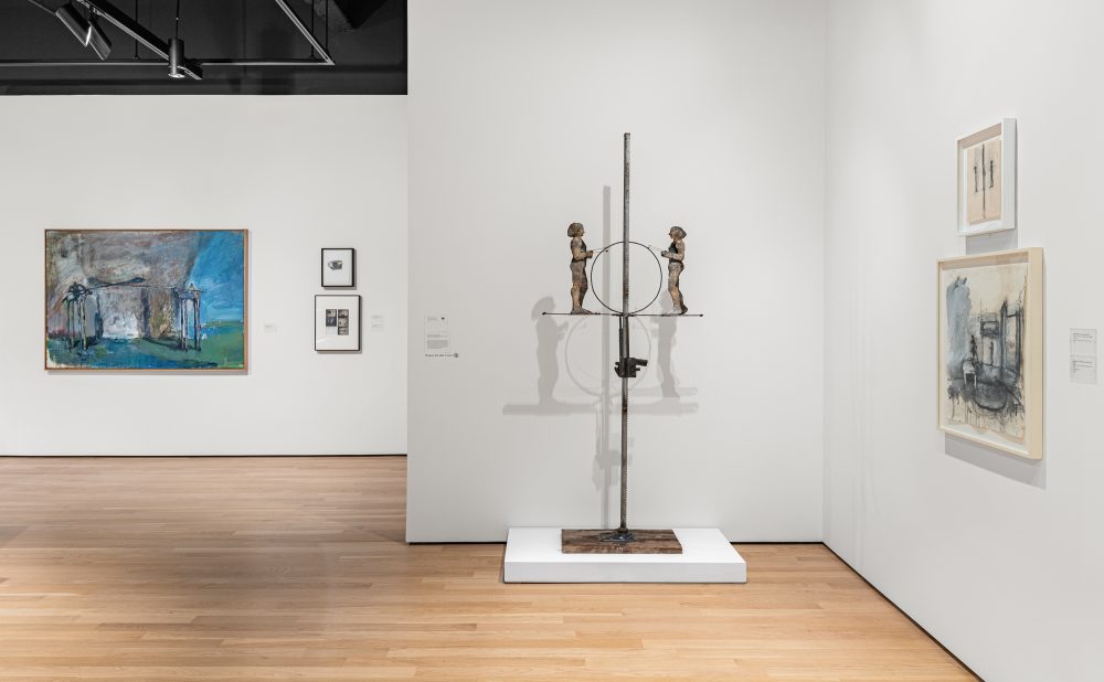 Art gallery with a sculpture representing two figures on a jack, and abstract paintings and drawings on white walls.