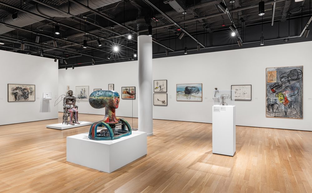 Art gallery with sculptures and paintings on display, featuring a colorful head-shaped sculpture on a low white pedestal and various abstract artworks on the wall.