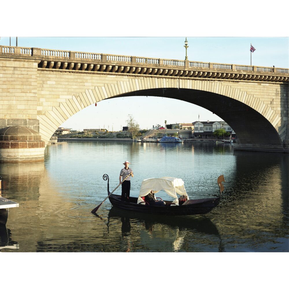 Color photograph of a stone arched bridge over water with a boat docked nearby under a clear sky. (Alt-text generated by AI)
