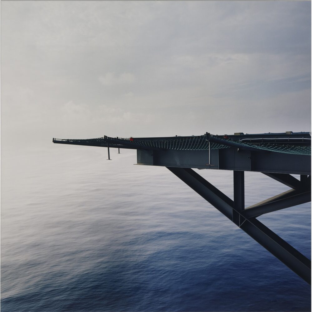 Perspective photograph looking along a dark metallic offshore platform extending over deep blue open water. (Alt-text generated by AI)