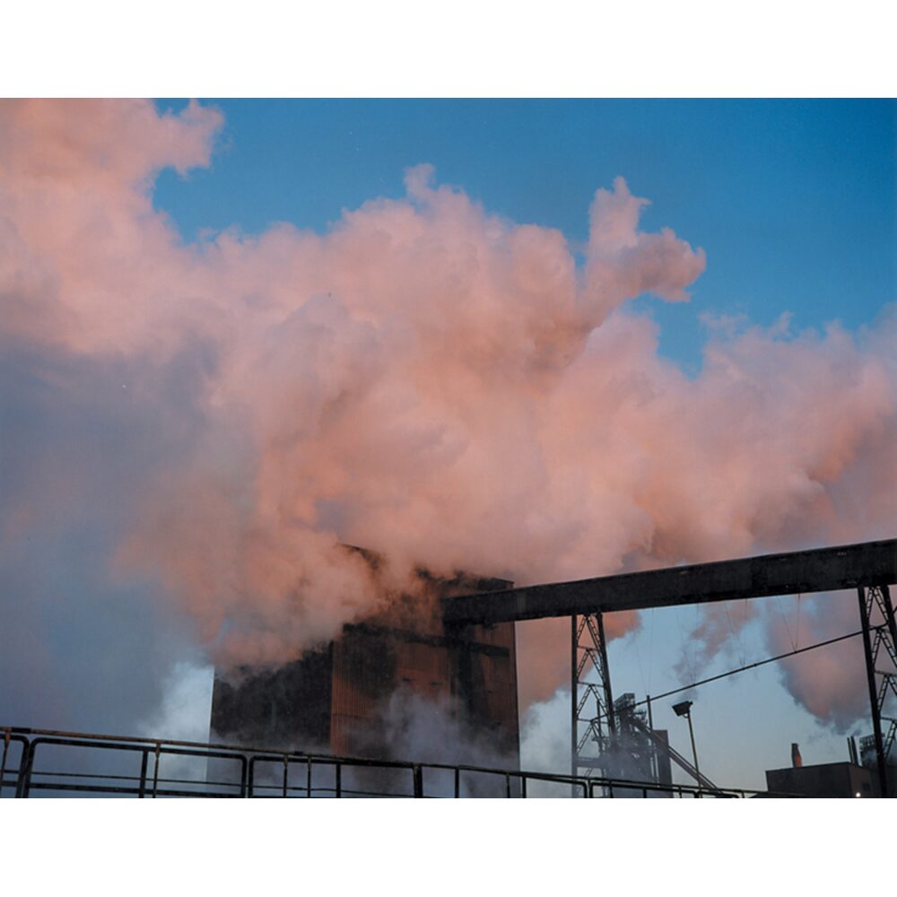 Industrial photograph showing thick, pink-tinted steam billowing from a factory structure against a blue sky. (Alt-text generated by AI)