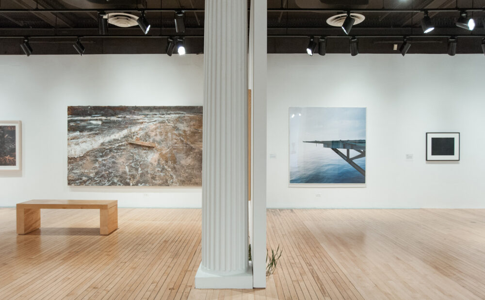 View of a gallery wall with a large landscape artwork on the left and a bench placed in front of it. (Alt-text generated by AI)