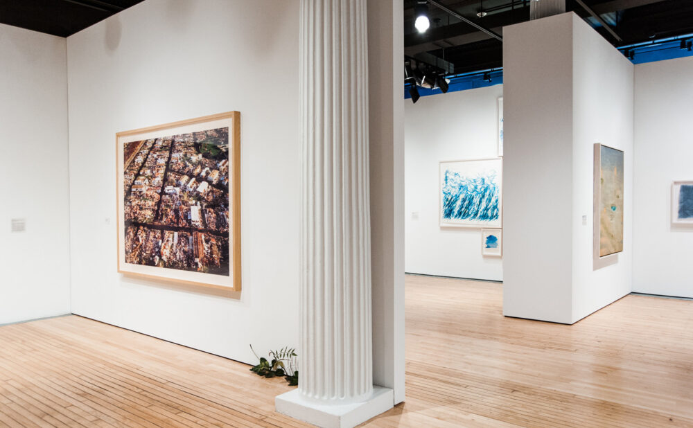 Gallery space with white architectural columns framing a framed artwork on the left wall and an open hallway beyond. (Alt-text generated by AI)
