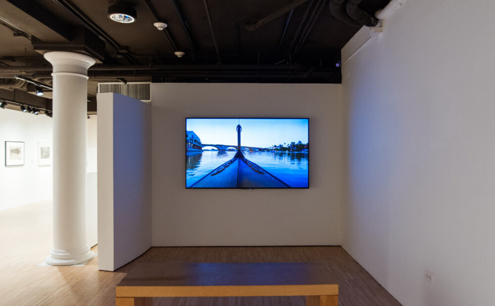 Media room in the gallery featuring a glowing blue video screen mounted on the wall above a viewing bench. (Alt-text generated by AI)