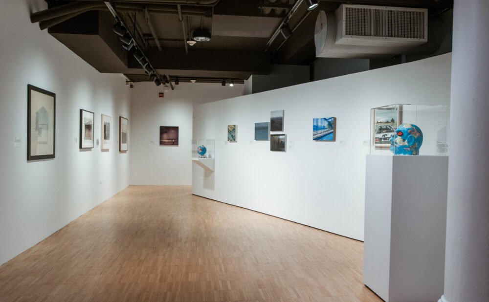 Perspective view down a gallery corridor leading past small framed artwork to a sculpture on a white pedestal. (Alt-text generated by AI)