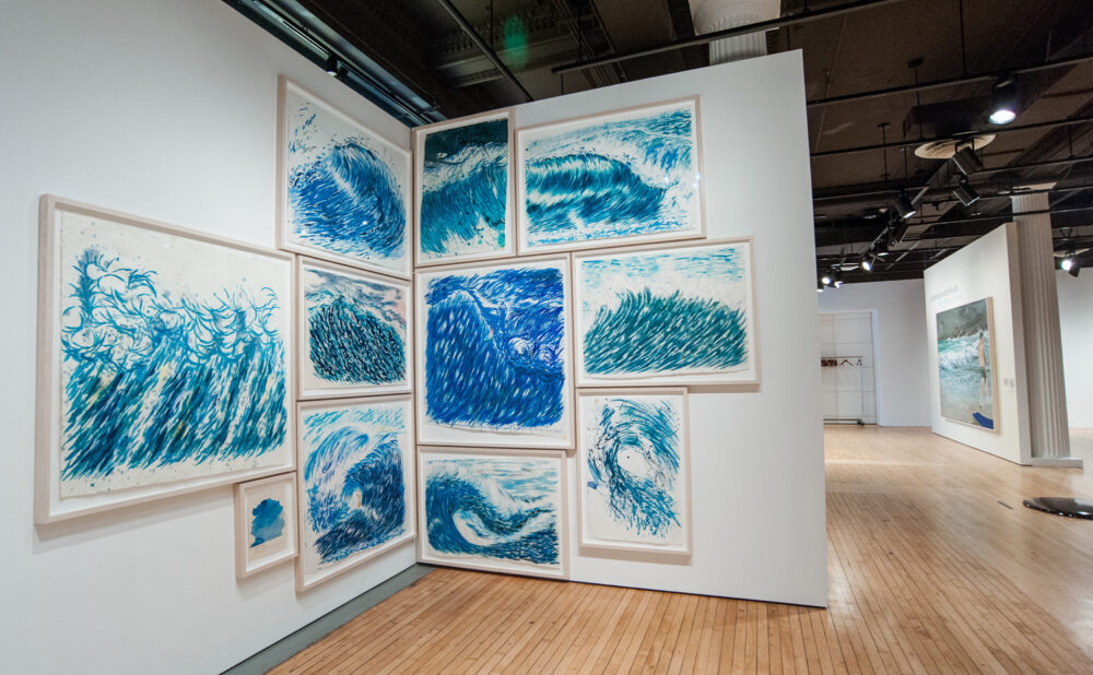 Gallery corner wall lined with blue-toned landscape drawings and sketches arranged in a loose grid layout. (Alt-text generated by AI)