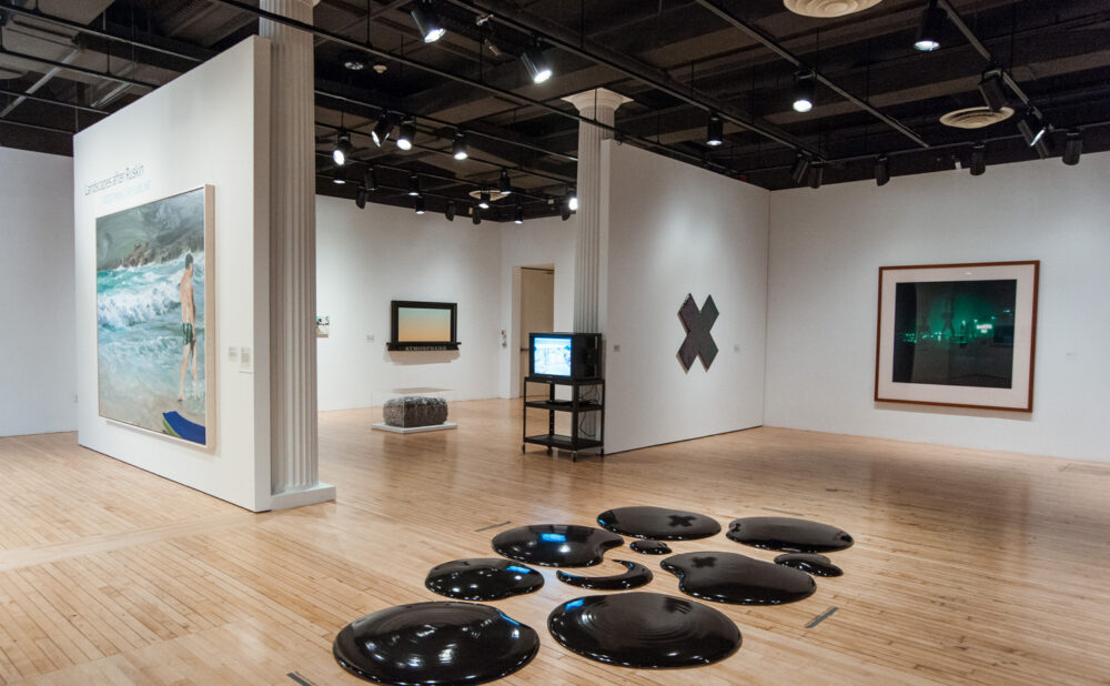 Wide gallery view showing dark oval floor sculptures arranged in the center of the hardwood floor. (Alt-text generated by AI)
