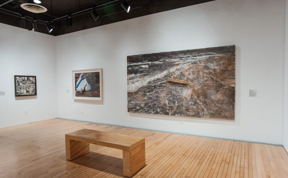 Exhibition wall displaying a wide landscape artwork above a simple wooden bench. (Alt-text generated by AI)