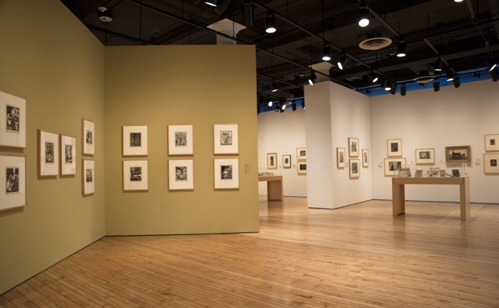 An exhibition corner showing multiple small, framed prints neatly arranged in rows on light tan walls around hardwood flooring. Alt text generated by AI