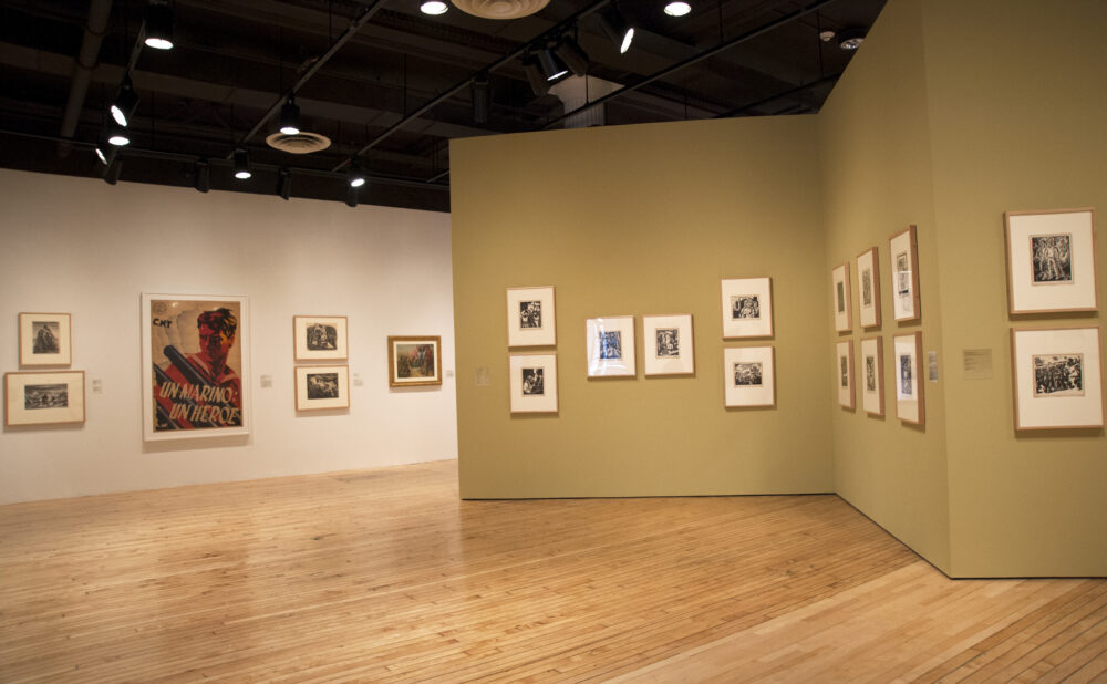 An exhibition corner showing multiple small, framed prints neatly arranged in rows on light tan walls around hardwood flooring. Alt text generated by AI