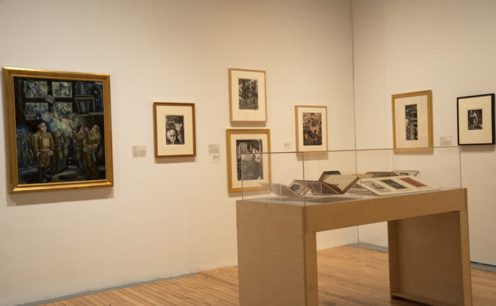 A gallery corner with a painting, framed prints, and a wooden display table set on light hardwood floors. Alt text generated by AI