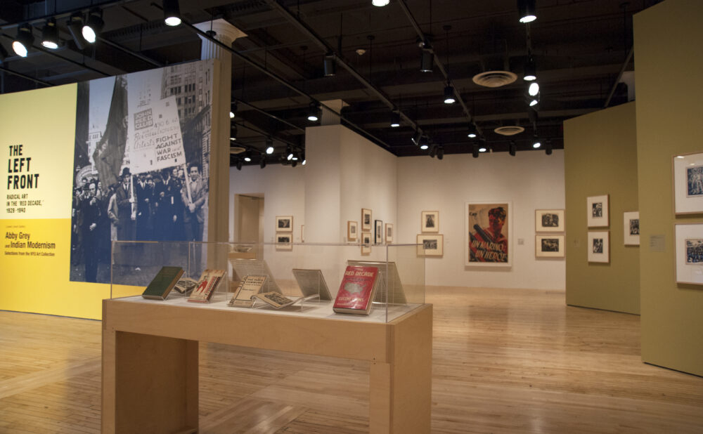 A wide view of an art gallery space with light wood floors, beige walls, and display cases showcasing artwork and books. Alt text generated by AI