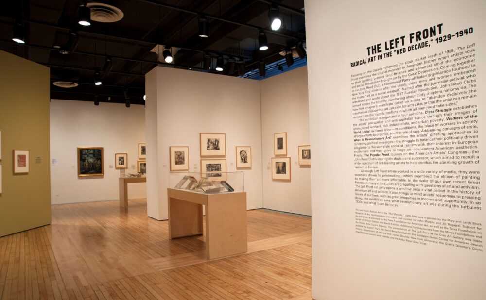 An art gallery exhibition room with a large text panel on a foreground wall leading into an open room with artwork displays and wooden pedestals. Alt text generated by AI