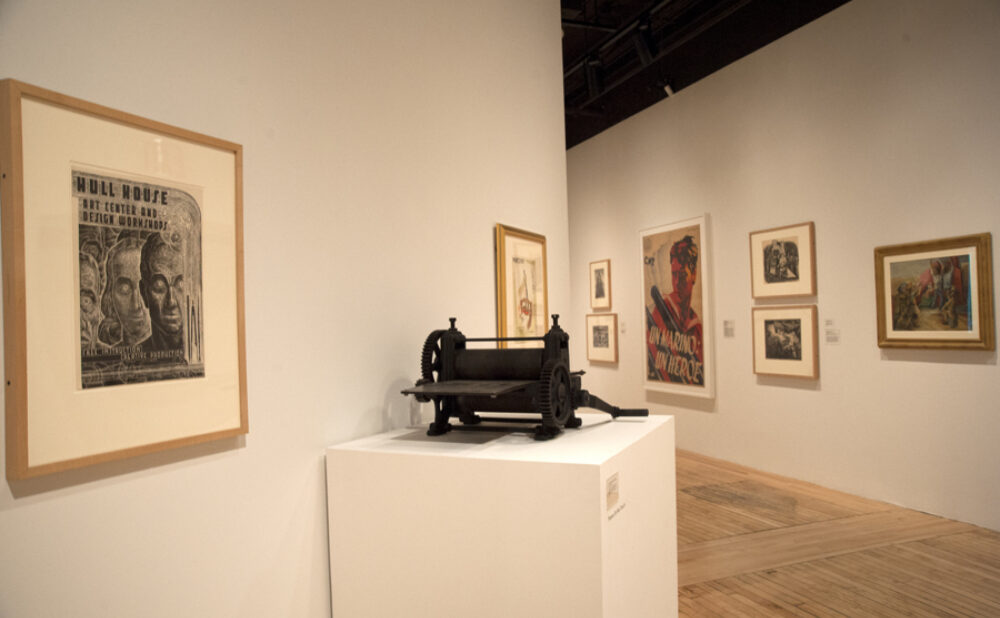 A gallery perspective highlighting framed prints on the wall and a dark mechanical printing press mounted on a central white pedestal. Alt text generated by AI