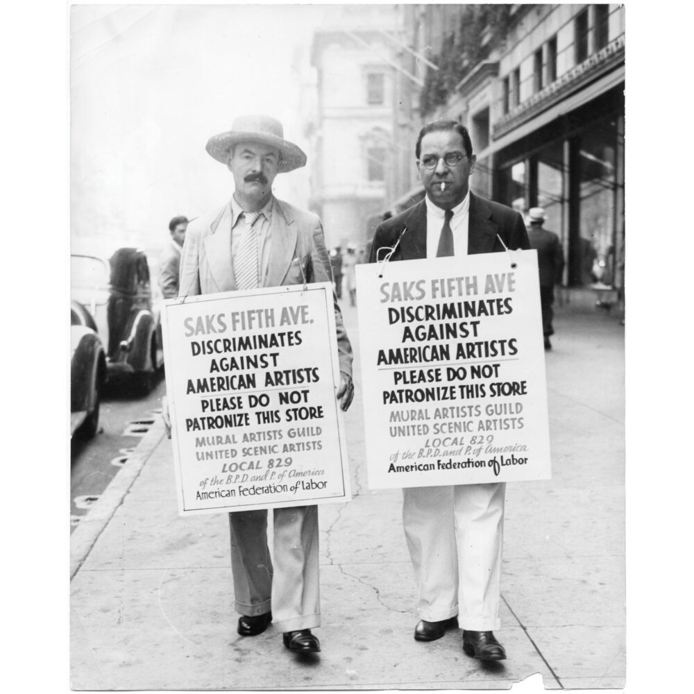 A black-and-white photograph of two men standing outdoors on a sidewalk, both wearing hats and holding large protest sandwich signs hanging from their shoulders. Alt text generated by AI
