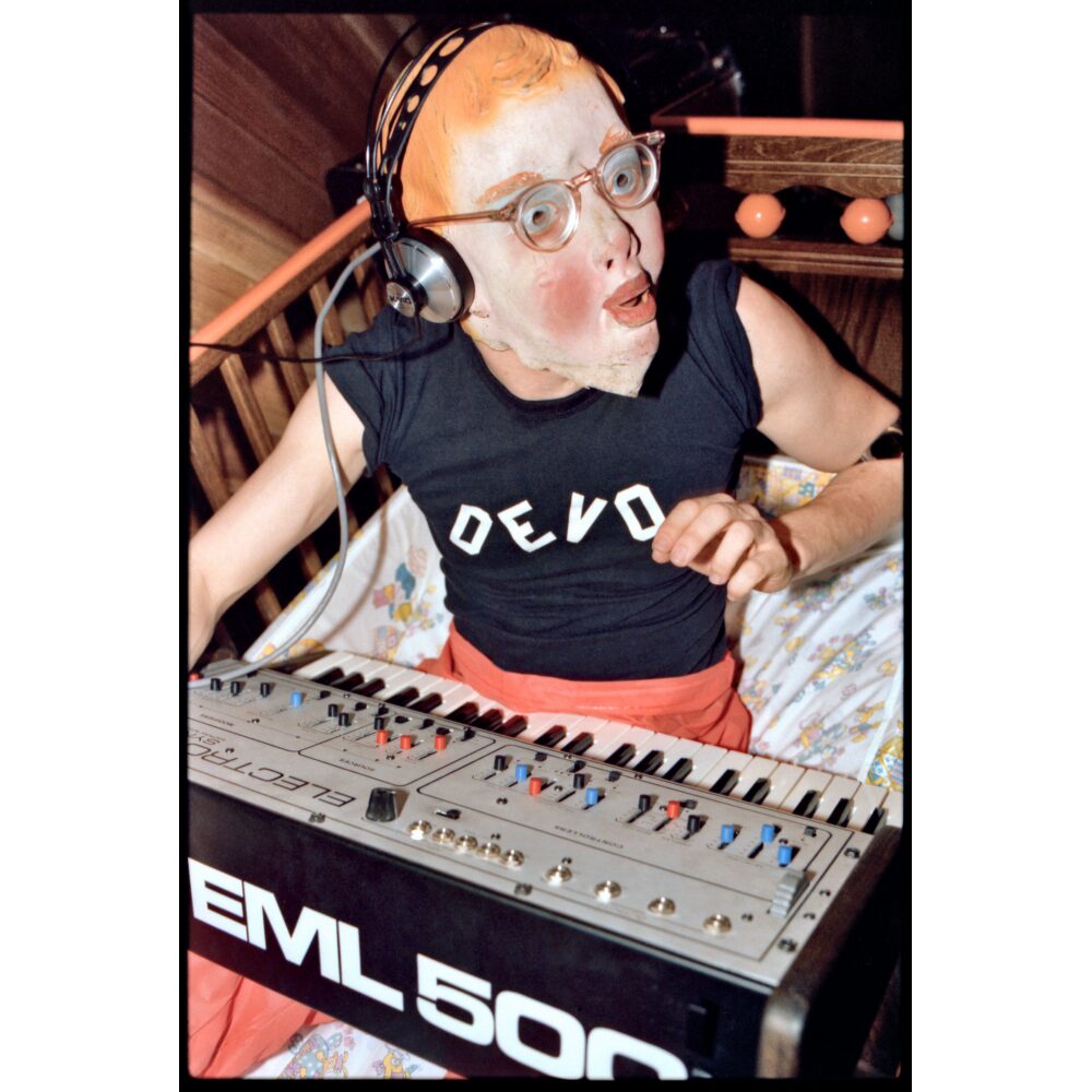 Photograph of Mark Mothersbaugh performing as "Booji Boy" in a baby mask, orange cap, and DEVO shirt behind a synthesizer. (Alt-text generated by AI)